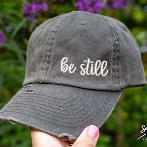 May include: A distressed gray baseball cap with the embroidered phrase "be still" in white script. The cap has a curved brim and a worn appearance, suggesting a vintage or casual style.