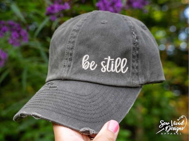 May include: A distressed gray baseball cap with the embroidered phrase "be still" in white script. The cap has a curved brim and a worn appearance, suggesting a vintage or casual style.