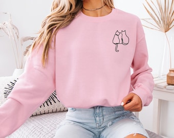 Peut inclure: Un sweat-shirt rose avec un dessin simple de deux chats assis dos à dos sur le devant.