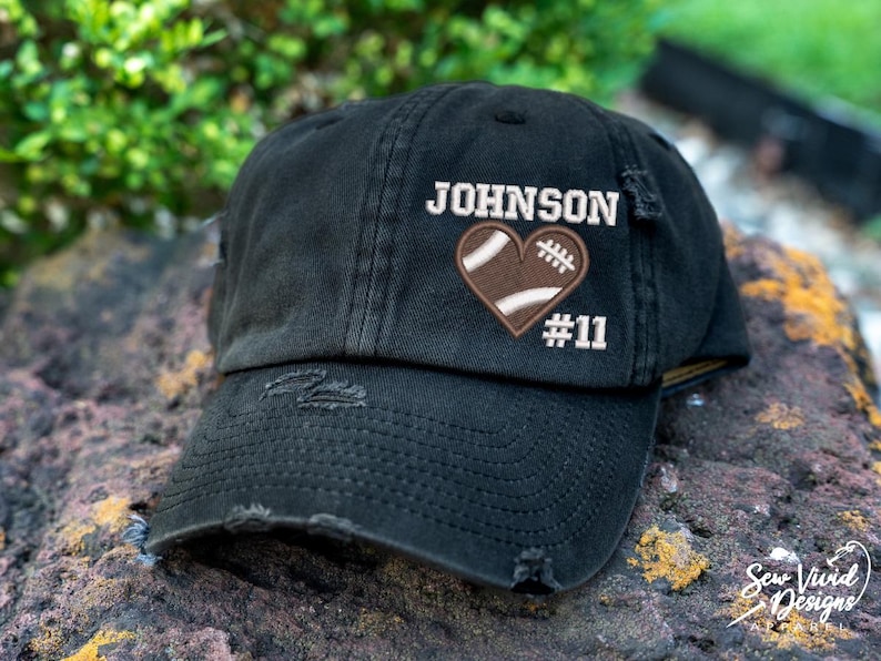 May include: Black distressed baseball cap with a white embroidered heart featuring a football design and the text "JOHNSON #11".