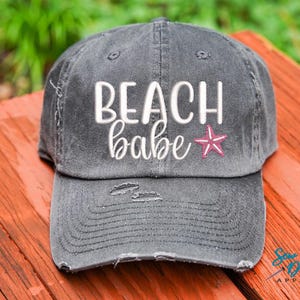 May include: A gray distressed baseball cap with the text "Beach babe" in white embroidery and a pink starfish.