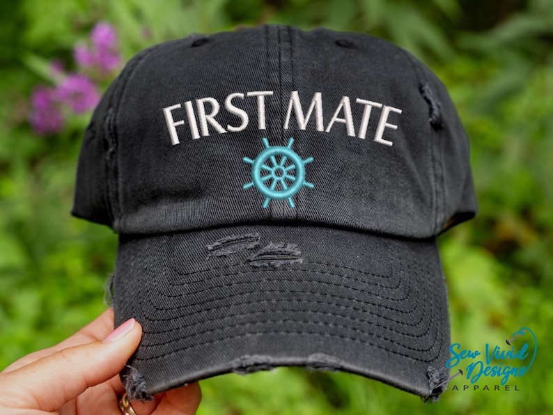 K&ouml;nnte beinhalten: Schwarze, ausgewaschene Baseballkappe mit dem gestickten Text "FIRST MATE" in Wei&szlig;, &uuml;ber einem t&uuml;rkisfarbenen Steuerraddesign. Die Kappe hat einen Used-Look mit abgenutzten Details.