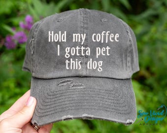Może przedstawiać: Szara, zniszczona czapka bejsbolowa z białym haftem "Hold my coffee I gotta pet this dog". Czapka ma wygląd vintage z przetarciami i zakrzywionym daszkiem. Tło to rozmyte zielone liście.