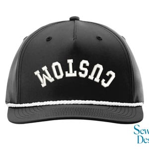 Può includere: Cappellino da baseball nero con la scritta "CUSTOM" in bianco, a lettere arcuate. Il cappellino ha un dettaglio di corda bianca lungo il bordo e un piccolo bottone nero sulla parte superiore. Il logo "Sew Vivid Designs" è in basso a destra.