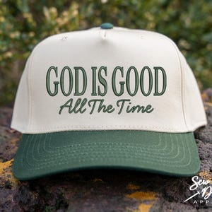 K&ouml;nnte beinhalten: Eine cremefarbene Baseballkappe mit dunkelgr&uuml;nem Schirm. Auf der Vorderseite befindet sich eine dunkelgr&uuml;ne Stickerei mit dem Text "GOD IS GOOD" in fetter Gro&szlig;schrift, darunter steht "All The Time" in einer kleineren, kursiven Schriftart.