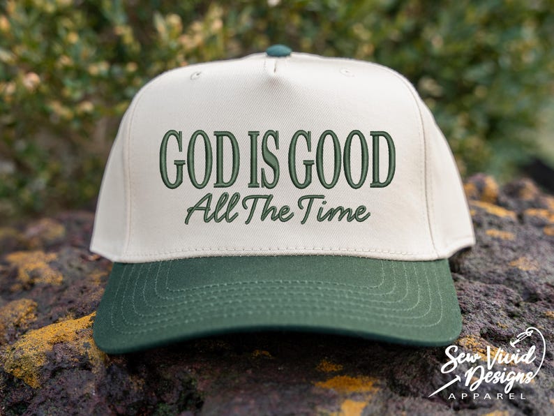 K&ouml;nnte beinhalten: Eine cremefarbene Baseballkappe mit dunkelgr&uuml;nem Schirm. Auf der Vorderseite befindet sich eine dunkelgr&uuml;ne Stickerei mit dem Text "GOD IS GOOD" in fetter Gro&szlig;schrift, darunter steht "All The Time" in einer kleineren, kursiven Schriftart.