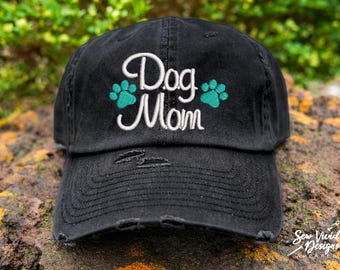 May include: Black distressed baseball cap with white embroidery that reads "Dog Mom" and two teal paw prints.