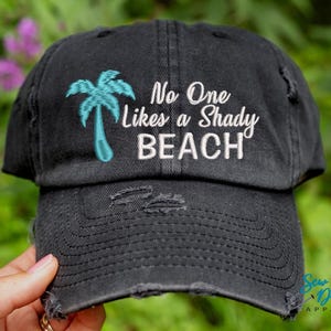 May include: Black distressed baseball cap with white embroidery that reads "No One Likes a Shady Beach". A turquoise palm tree is embroidered on the left side of the cap.