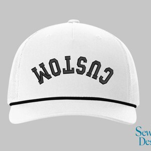 May include: White trucker hat with the word "CUSTOM" embroidered in black block letters. The hat has a black rope detail and a mesh back. The Sew Vivid Designs logo is in the bottom right corner.