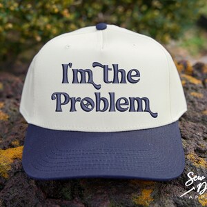 May include: A baseball cap with a cream-colored crown and a navy blue brim. The phrase "I'm the Problem" is embroidered in navy blue script across the front. The cap is sitting on a rock.