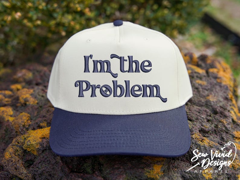 May include: A baseball cap with a cream-colored crown and a navy blue brim. The phrase "I'm the Problem" is embroidered in navy blue script across the front. The cap is sitting on a rock.