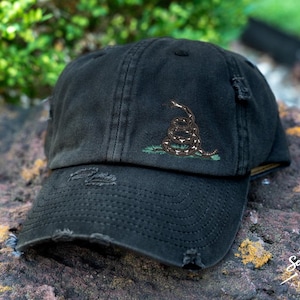 May include: A black distressed baseball cap with a green and brown embroidered snake design. The snake is coiled and has a white stripe down its back. The cap has a distressed look with holes and frayed edges.
