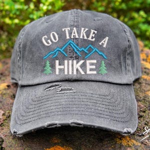 Op de afbeelding: Een grijze, verweerde baseballpet met de tekst "Go Take A Hike" geborduurd in wit. De borduurwerk bevat ook blauwe bergen en groene bomen.