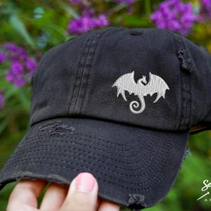 May include: A black distressed baseball cap with a white embroidered dragon design. The cap has a curved brim and a distressed look.