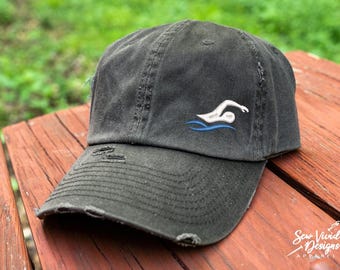 May include: A black distressed baseball cap with a white embroidered swimmer silhouette and blue water design. The cap has a curved brim and a distressed look.