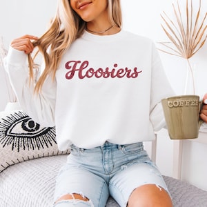 May include: White sweatshirt with the word "Hoosiers" in a script font. The text is in a maroon color. A person is holding a green mug that says "Coffee". The person is wearing blue denim shorts.