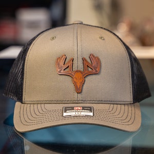 May include: A khaki and black trucker hat with a brown leather deer skull and antlers patch. The hat has a mesh back and a curved peak. The tag on the front reads "RICHARDSON 112".