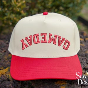 May include: A cream-colored baseball cap with a red brim and a red button on top. The front features the word GAMEDAY embroidered in red block letters, positioned in an upside-down arc across the center of the crown of the hat.