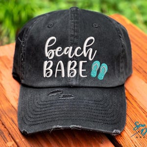 May include: Black distressed baseball cap with white embroidery that reads "beach babe" and turquoise blue flip-flop icons.