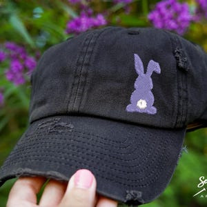 May include: A black distressed baseball cap with a purple embroidered bunny rabbit on the front. The cap has a curved brim and a sewn-on adjustable strap.