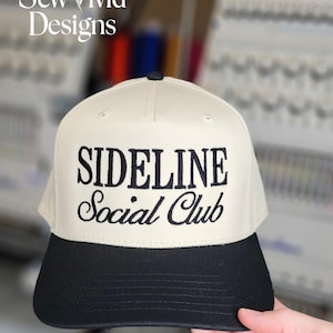 May include: A beige and black baseball cap with the words "SIDELINE Social Club" embroidered in black. The cap has a curved brim and a structured crown. The logo "Sew Vivid Designs" is in the upper left corner.