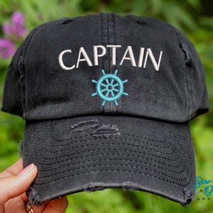 K&ouml;nnte beinhalten: Schwarze Baseballkappe im Used-Look mit dem Wort "CAPTAIN" in Wei&szlig; gestickt und einem t&uuml;rkisfarbenen Steuerraddesign. Die Kappe hat einen vorgewaschenen Look mit ausgefransten Kanten, ein Accessoire im maritimen Stil.