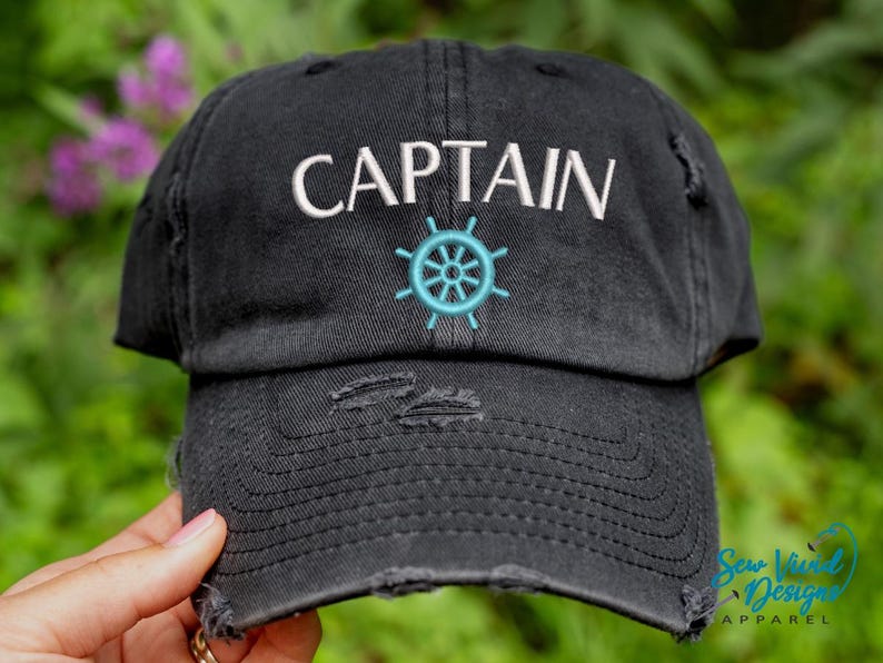 K&ouml;nnte beinhalten: Schwarze Baseballkappe im Used-Look mit dem Wort "CAPTAIN" in Wei&szlig; gestickt und einem t&uuml;rkisfarbenen Steuerraddesign. Die Kappe hat einen vorgewaschenen Look mit ausgefransten Kanten, ein Accessoire im maritimen Stil.