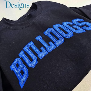 May include: A black crewneck sweatshirt featuring the word BULLDOGS in large, blue, glittery, embroidered block letters across the chest. The top left corner displays the text Sew Vivid Designs in a blue serif font on a white background.