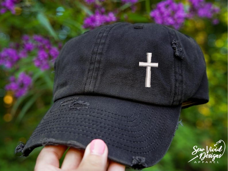 Op de afbeelding: Zwarte, distressed baseballcap met een wit geborduurd kruis op de voorkant. De cap heeft een gebogen klep en een versleten uitstraling. Paarse bloemen zijn zichtbaar op de achtergrond.