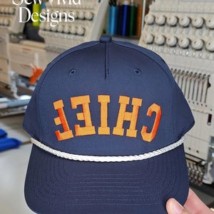 Puede incluir: Gorra de b&eacute;isbol azul marino con la palabra "CHIEF" bordada en naranja y rojo. La gorra tiene un detalle de cuerda blanca a lo largo del borde. El texto "Sew Vivid Designs" est&aacute; en la esquina superior izquierda.