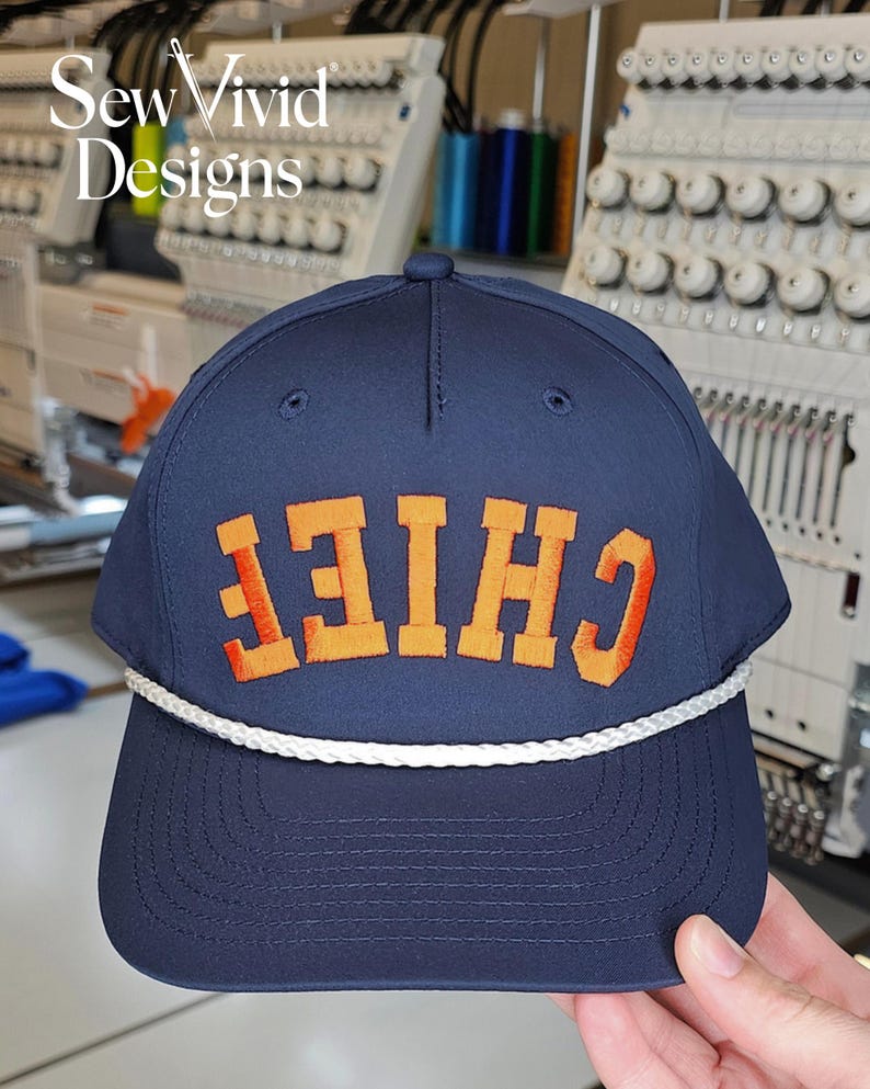 Puede incluir: Gorra de b&eacute;isbol azul marino con la palabra "CHIEF" bordada en naranja y rojo. La gorra tiene un detalle de cuerda blanca a lo largo del borde. El texto "Sew Vivid Designs" est&aacute; en la esquina superior izquierda.