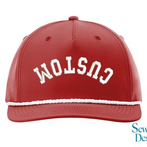 Puede incluir: Gorra de b&eacute;isbol roja con la palabra "CUSTOM" bordada en blanco, arqueada en la parte delantera. La gorra tiene un detalle de cuerda blanca a lo largo del borde y un peque&ntilde;o bot&oacute;n en la parte superior. El logotipo de Sew Vivid Designs est&aacute; en la esquina inferior derecha.