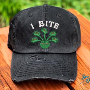 May include: Black distressed baseball cap with the embroidered phrase "I BITE" above a green Venus flytrap design. The cap has a curved brim and a worn appearance, suggesting a vintage or casual style.