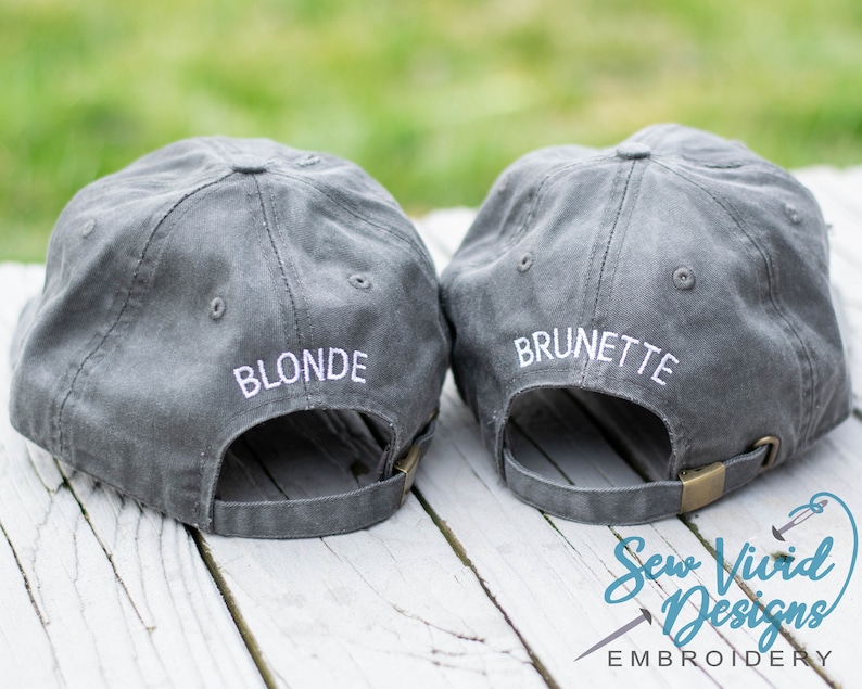 May include: Two gray baseball caps with white embroidered text. The cap on the left says "Blonde" and the cap on the right says "Brunette".