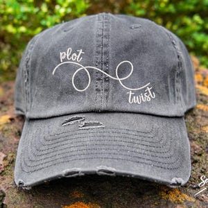 May include: A black distressed baseball cap with a white embroidered design that says "plot twist" with a stylized infinity symbol.