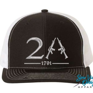May include: Black and white trucker hat with a white mesh back. The front of the hat features a silver embroidered design of the number 2A with two rifles and the text "1791".