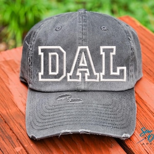 Może przedstawiać: Szara, zniszczona czapka bejsbolówka z białym haftem "DAL" z przodu.
