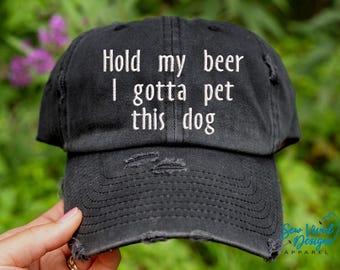 May include: Black distressed baseball cap with white embroidered text that reads "Hold my beer I gotta pet this dog".