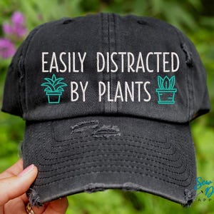 May include: Black distressed baseball cap with white text that reads "Easily Distracted by Plants". The text is surrounded by two green plant illustrations.
