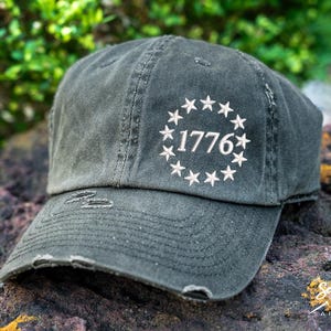 May include: A black distressed baseball cap with a white circle embroidered with 13 stars and the year "1776" in the center.
