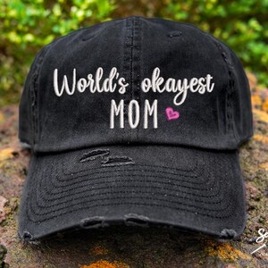 May include: Black distressed baseball cap with white embroidered text that reads "World's okayest MOM" with a pink heart.