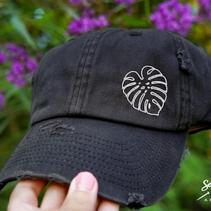 Puede incluir: Una gorra de b&eacute;isbol negra desgastada con un dise&ntilde;o de hoja de monstera bordada en blanco. La gorra tiene una visera curvada y una etiqueta cosida que dice "Sew Vivid Designs Apparel".