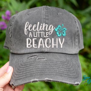 May include: A gray distressed baseball cap with the text "feeling a little beachy" embroidered in white and a turquoise hibiscus flower embroidered on the front.
