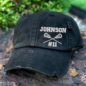 May include: Black distressed baseball cap with white embroidered text "JOHNSON #11" and two lacrosse sticks.