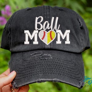 May include: Black baseball cap with distressed detailing. The cap features white embroidery that reads "Ball Mom" with a heart-shaped softball graphic.