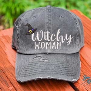 May include: A gray distressed baseball cap with the text "Witchy Woman" embroidered in white. A black witch's hat with a purple band is embroidered above the text.