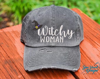 May include: A gray distressed baseball cap with the text "Witchy Woman" embroidered in white. A black witch's hat with a purple band is embroidered above the text.