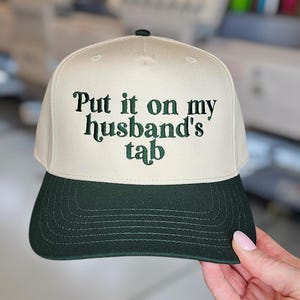 May include: A baseball cap with a cream-colored crown and a dark green brim. The text "Put it on my husband's tab" is embroidered in dark green on the front of the cap. The cap is a classic six-panel design.