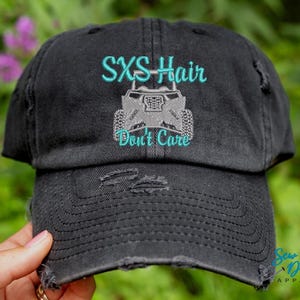 Może przedstawiać: Czarna, zniszczona czapka bejsbolówka z turkusowym haftem, na którym widnieje napis "SXS Hair Don't Care" oraz szary haft przedstawiający pojazd typu side-by-side.