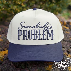 May include: A baseball cap with a cream-coloured crown and a navy blue brim. The phrase "Somebody's PROBLEM" is embroidered in navy blue script across the front. The cap has a curved brim and a button on top. The Sew Vivid Designs Apparel logo is on the bottom right.